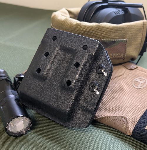 A black Kydex magazine holder with multiple mounting holes, resting on tactical gear. Surrounding the holder are a black flashlight, a pair of electronic hearing protection earmuffs with a tan cover, and a tan tactical glove with a logo. The background features a green fabric surface.
