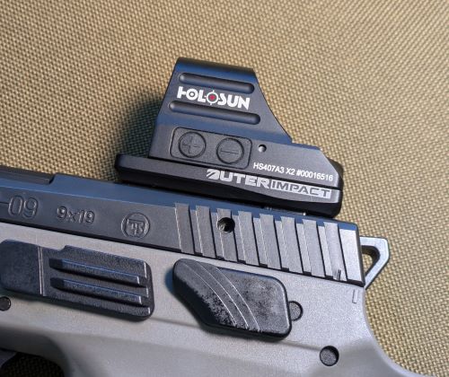 A close-up image of a pistol with a Holosun HS407A3 X2 red dot sight mounted on an OuterImpact adapter plate. The slide of the pistol has serrations, and the frame features a textured grip and an accessory rail. The image is set against a neutral fabric background.