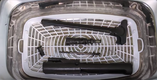 Dirty gun parts, including a bolt carrier group and charging handle, submerged in an ultrasonic cleaner with a white basket.